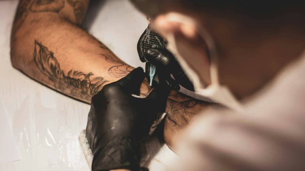 Tatuador con mascarilla y guantes trabajando sobre el antebrazo de un cliente