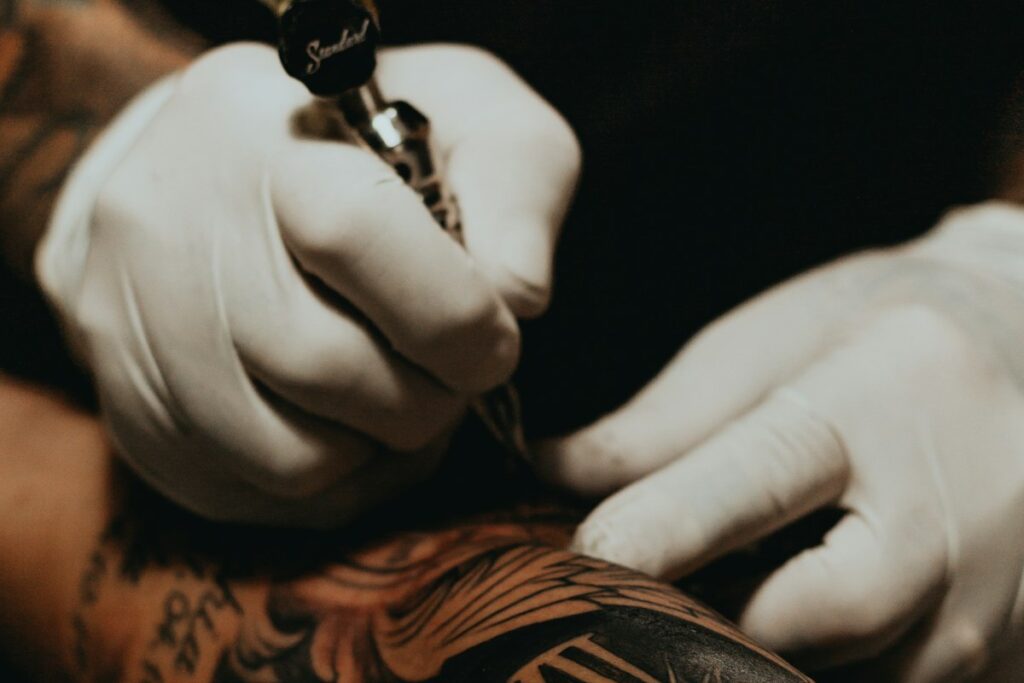 Manos con guantes blancos sujetando la máquina de tatuar sobre un brazo tatuado